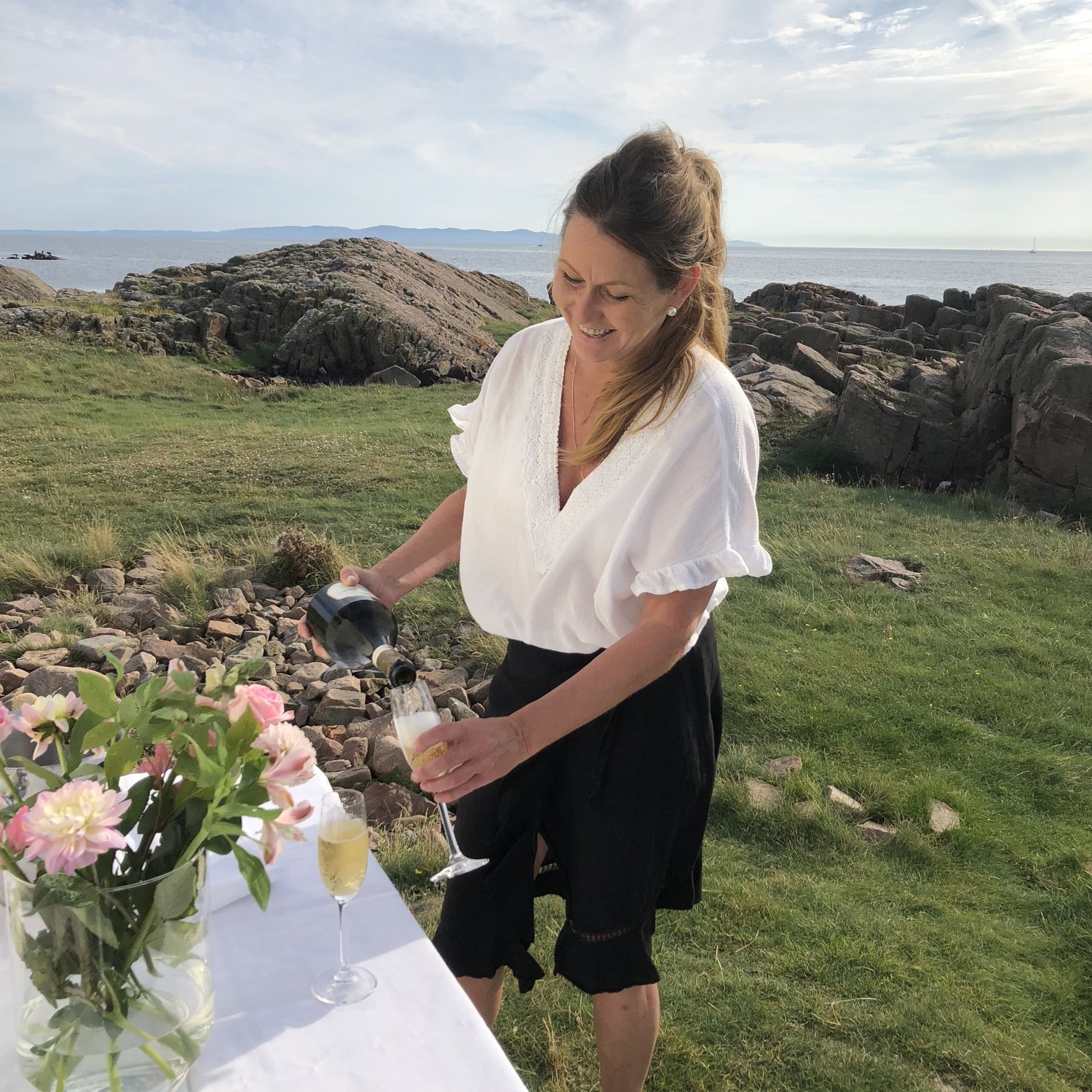 En kvinna som fyller ett glas med champagne vid ett utebord med blommor.
